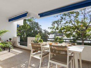 A balcony with a white table and chairs, a small potted plant, and a view of trees and buildings. - 2 Dormitorios en Medina Garden Ref 511 (Marbella)
