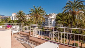 Una terraza en la azotea con mobiliario exterior blanco, palmeras y vistas a edificios al fondo.