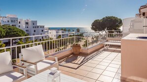 Una terraza con mobiliario blanco, vista al mar y edificios al fondo.