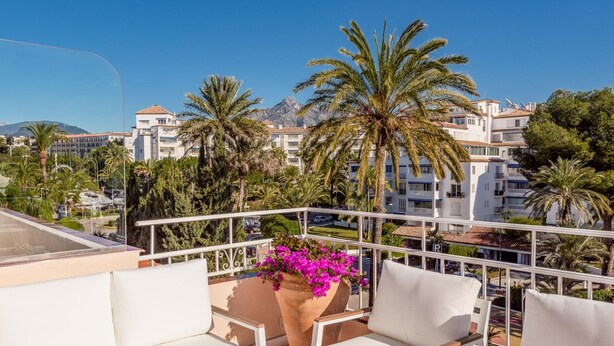 Un balcón con mobiliario exterior blanco, una planta en maceta con flores rosadas y vistas a palmeras y edificios.