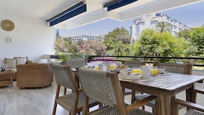 A balcony with wicker furniture, a table set for breakfast, and a view of buildings and greenery. - 2 Dormitorios en Medina Garden Ref 711 (Marbella)