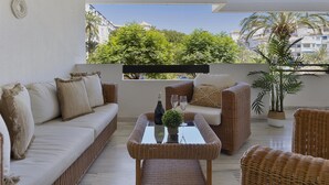 A balcony with a wicker sofa, a glass-top table with a bottle and glasses, and a potted plant. - 2 Dormitorios en Medina Garden Ref 711 (Marbella)