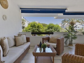 A balcony with a wicker sofa, a glass-top table with a bottle and glasses, and a potted plant. - 2 Dormitorios en Medina Garden Ref 711 (Marbella)