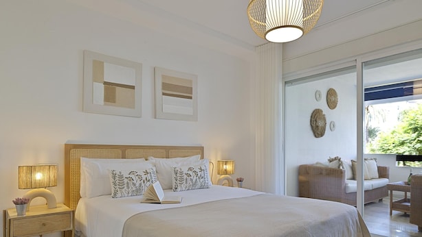 A bedroom with a large bed, bedside lamps, a nightstand, and a view of an outdoor seating area. - 2 Dormitorios en Medina Garden Ref 711 (Marbella)