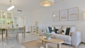 A modern living room with a sofa, coffee table, and dining area with chairs and a table. - 2 Dormitorios en Medina Garden Ref 711 (Marbella)