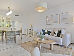 A modern living room with a sofa, coffee table, and dining area with chairs and a table. - 2 Dormitorios en Medina Garden Ref 711 (Marbella)