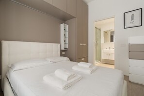 Quarto de hotel com uma cama, um guarda-roupa branco, um banheiro visível através de uma porta de vidro e um quadro na parede.