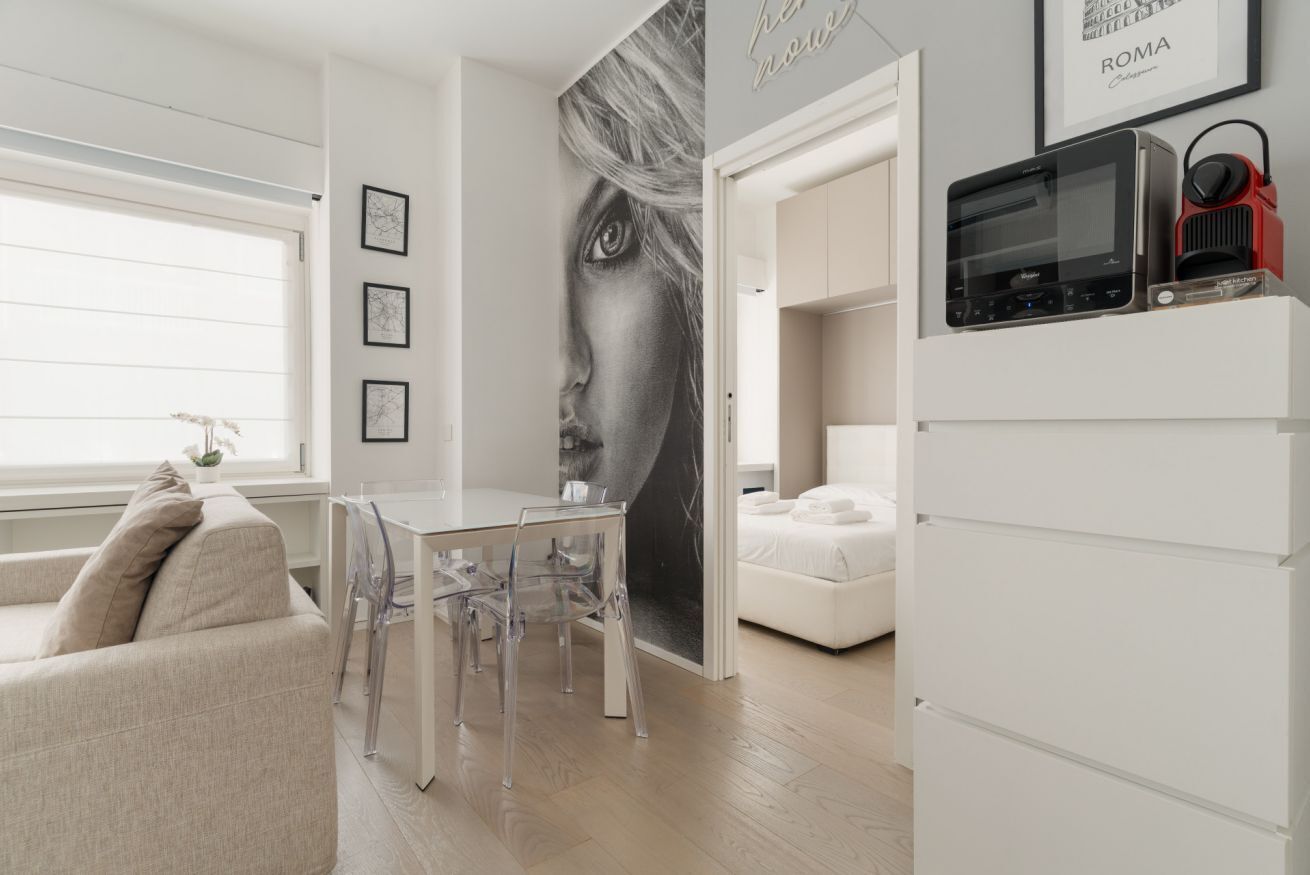 A modern living room with a dining area, a white sofa, a glass table, and chairs. There is a large wall mural, a white bed, and a white cabinet with a radio.