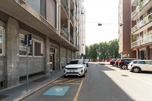 Vista da rua com um carro branco estacionado na lateral, uma vaga de estacionamento para deficientes azuis e edifícios com varandas.