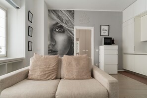 A living room with a beige sofa, a large wall mural, and a kitchen area with white cabinets. - GuestHost - Cozy Apartment in Darsena, Near the Navigli (Milan)