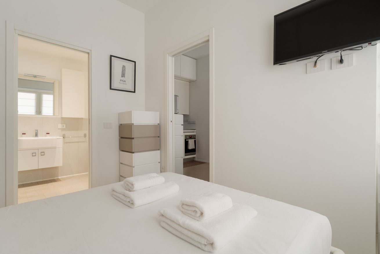 A hotel room with a bed, a flat-screen TV mounted on the wall, a framed picture on the wall, and a bathroom visible through an open door.