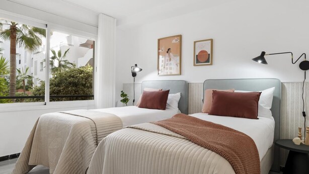 Quarto de hotel moderno com duas camas, uma janela grande com vista para palmeiras e uma pequena mesa de cabeceira com abajur.