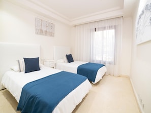 Ein Hotelzimmer mit zwei Betten, einem Nachttisch, einem Fenster mit durchsichtigen Vorhängen und einem an der Wand hängenden Kunstwerk.
