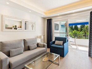 Ein modernes Wohnzimmer mit Sofa, Sesseln, einem Glastisch und einem Balkon mit Blick auf Grünflächen.