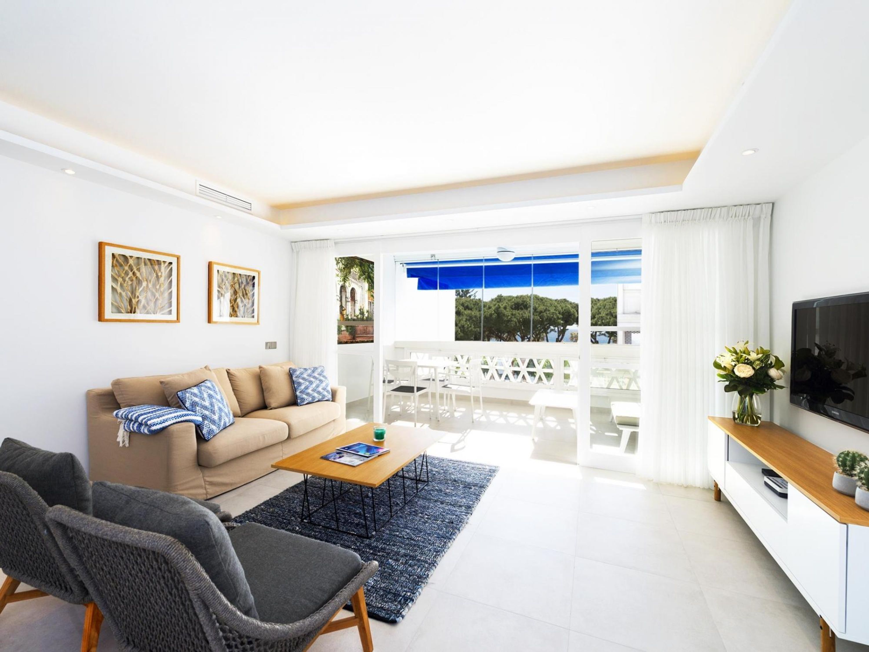 Una sala de estar moderna con un sofá, una mesa de centro y sillas, un televisor de pantalla plana, y vistas al exterior.
