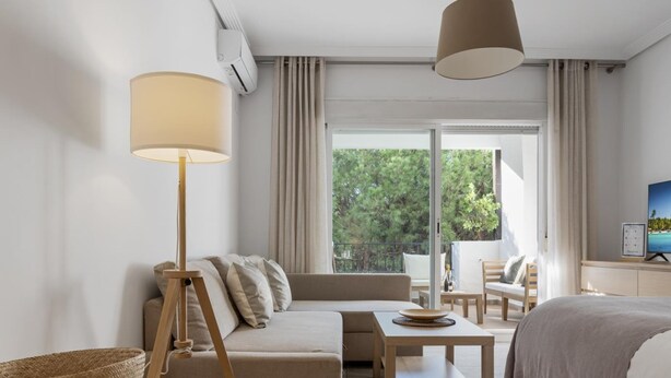 Een moderne hotelkamer met een bank, een bed, een eettafel en een groot raam met uitzicht op bomen.