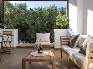 A balcony with a wooden table, two chairs, a sofa, and a bottle of champagne. - Confortable Estudio en Medina Garden ref 332 (Marbella)