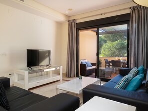 Ein modernes Wohnzimmer mit einem Flachbildfernseher auf einem Glastisch, einer Couch und einem Balkon mit Gartenmöbeln.