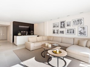 Ein modernes Wohnzimmer mit einem beigefarbenen Ecksofa, einem runden Couchtisch mit Kerzen und einer Küchenzeile mit dunklen Schränken.