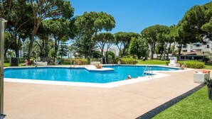 A swimming pool with a clear blue water, surrounded by trees and a building in the background. - 2 Dormitorios Playas del Duque Ref 228 (Marbella)