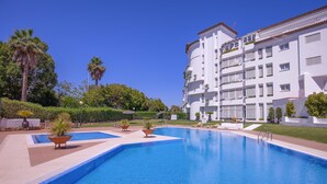A swimming pool with a clear blue water surface, surrounded by a white building with balconies and a garden area with palm trees and shrubs. - Elegante Apartamento en Las Gaviotas (Marbella)