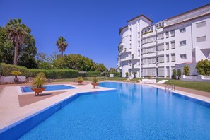A swimming pool with a clear blue water surface, surrounded by a white building with balconies and a garden area with palm trees and shrubs. - Elegante Apartamento en Las Gaviotas (Marbella)