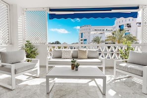 A balcony with a white outdoor seating area, a table, and a view of a building with the sign 'VAS-DUQUE'. - Elegante Apartamento en Las Gaviotas (Marbella)