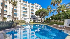 A swimming pool with clear blue water, surrounded by a well-maintained garden with palm trees and a multi-story hotel building in the background. - Elegante Apartamento en Las Gaviotas (Marbella)