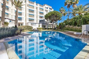 A swimming pool with clear blue water, surrounded by a well-maintained garden with palm trees and a multi-story hotel building in the background. - Elegante Apartamento en Las Gaviotas (Marbella)