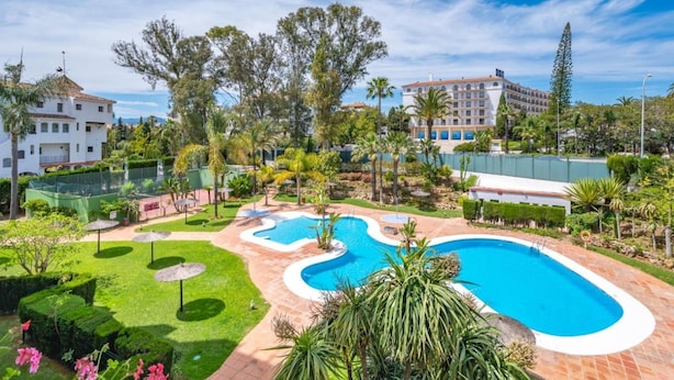Een resort met een groot zwembad, weelderige begroeiing en een helderblauwe lucht.