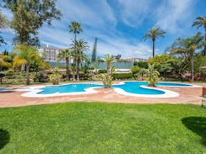 A pool area with a clear sky, surrounded by palm trees and a well-maintained lawn. - Estudio en Medina Garden Ref 362 (Marbella)