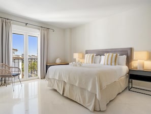 Ein Hotelzimmer mit einem großen Bett, zwei Nachttischen, einem Sessel und einem Balkon mit Vorhängen.