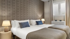 Et hotelværelse med to senge, et natbord med lampe, en kommode og et vindue med gardiner.