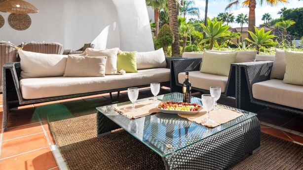 A sunlit outdoor seating area with a sofa and chairs, a glass coffee table with a fruit platter, and a bottle of wine. - Fant Stico Apartamento en Jardines de Ventura (Marbella)