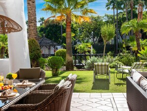 A patio with wicker furniture, a dining table set with plates and glasses, and a view of a lush garden with palm trees and a white building in the background. - Fant Stico Apartamento en Jardines de Ventura (Marbella)