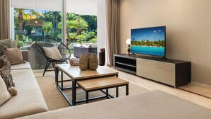 A modern living room with a sofa, a coffee table, a TV on a stand, and a view of a tropical garden through large windows. - Fant Stico Apartamento en Jardines de Ventura (Marbella)