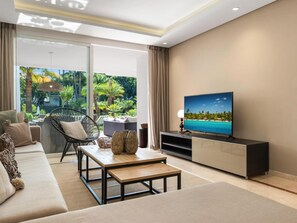 A modern living room with a sofa, a coffee table, a TV on a stand, and a view of a tropical garden through large windows. - Fant Stico Apartamento en Jardines de Ventura (Marbella)