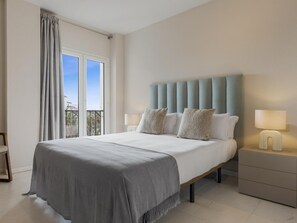 A hotel room with a large bed, a bedside table with a lamp, a view of the outdoors through a window, and a balcony. - Espacioso y Encantador Apartamento en Las Gaviotas (Marbella)