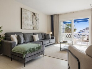 A modern living room with a grey sofa, a green throw, a floor lamp, and a balcony with a view of the sea. - Espacioso y Encantador Apartamento en Las Gaviotas (Marbella)