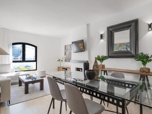 Ein modernes Wohnzimmer mit einem Glastisch, grauen Stühlen, einer Couch und einem Couchtisch, sowie einem an der Wand montierten Fernseher.