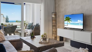 Un salon moderne avec un téléviseur diffusant une scène de plage, un canapé, une table basse avec une bouteille et des fleurs, et une vue sur l’extérieur.