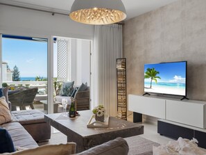 Ein modernes Wohnzimmer mit Fernseher, der eine Strandansicht zeigt, einer Couch, einem Couchtisch mit Flasche und Blumen sowie Blick ins Freie.