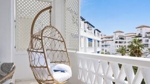 Un balcon doté d’un fauteuil suspendu en osier, d’une balustrade blanche en treillis et offrant une vue sur des bâtiments blancs aux toits recouverts de tuiles rouges.