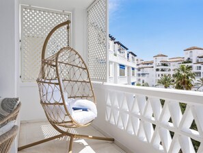 Ein Balkon mit einem Korbschaukelstuhl, einem weißen Gittergeländer und Blick auf weiße Gebäude mit roten Ziegeldächern.