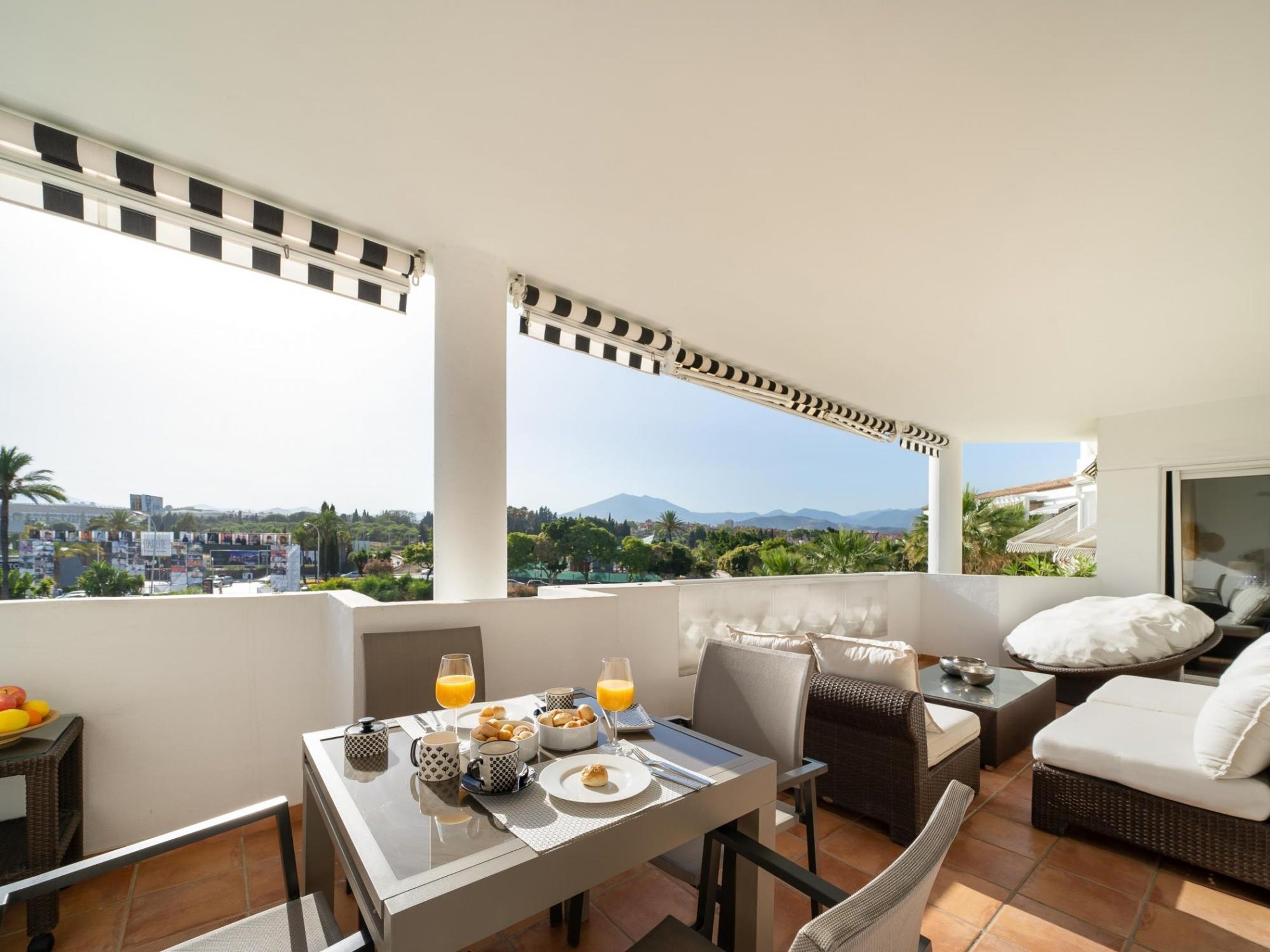 Ein Balkon mit Esstisch, gedecktem Frühstück, Korbmöbeln und Blick auf Stadt und Berge.
