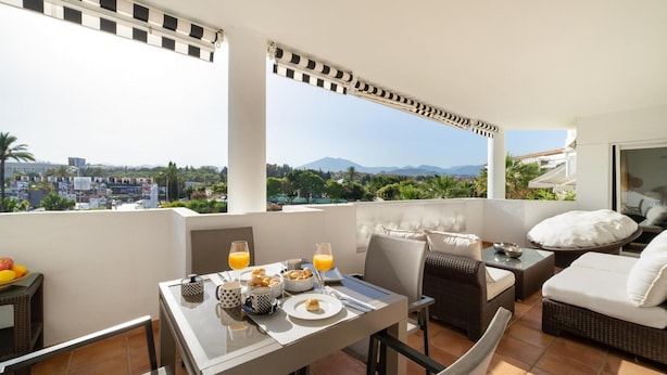A balcony with a dining table set for breakfast, wicker furniture, and a view of the city and mountains. - Apartamento en Playa Roc o (Marbella)