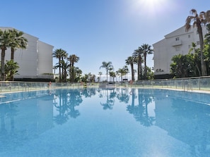 A clear blue pool surrounded by palm trees and white buildings. - Apartamento en Playa Roc o (Marbella)