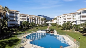 A swimming pool surrounded by a fence, with a clear sky and mountains in the background. - Apartamento en Playa Roc o (Marbella)
