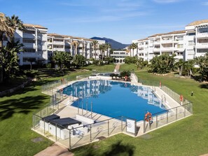 A swimming pool surrounded by a fence, with a clear sky and mountains in the background. - Apartamento en Playa Roc o (Marbella)