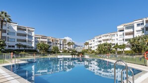 A swimming pool surrounded by multi-story buildings with balconies, clear skies, and palm trees. - Apartamento en Playa Roc o (Marbella)